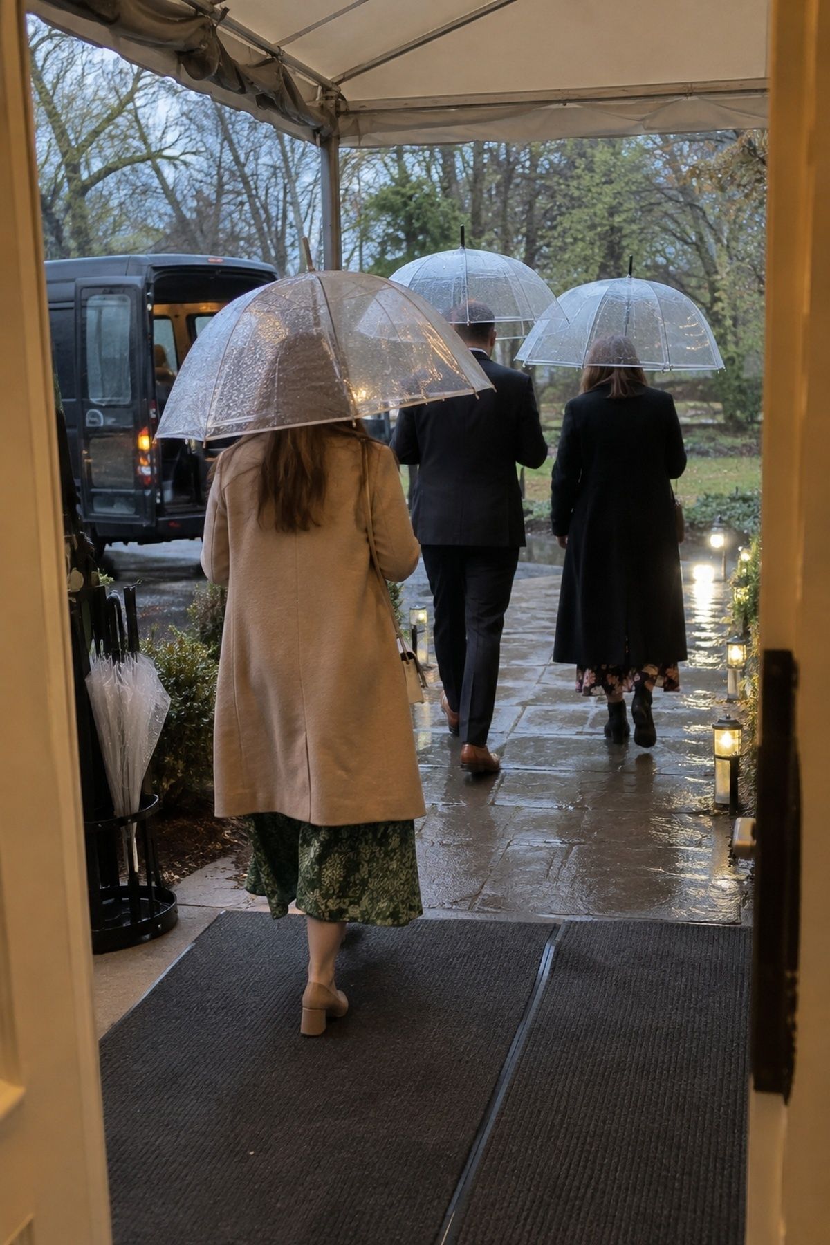 Wedding guests under clear umbrellas following a lit wet path from a shuttle to a covered entrance