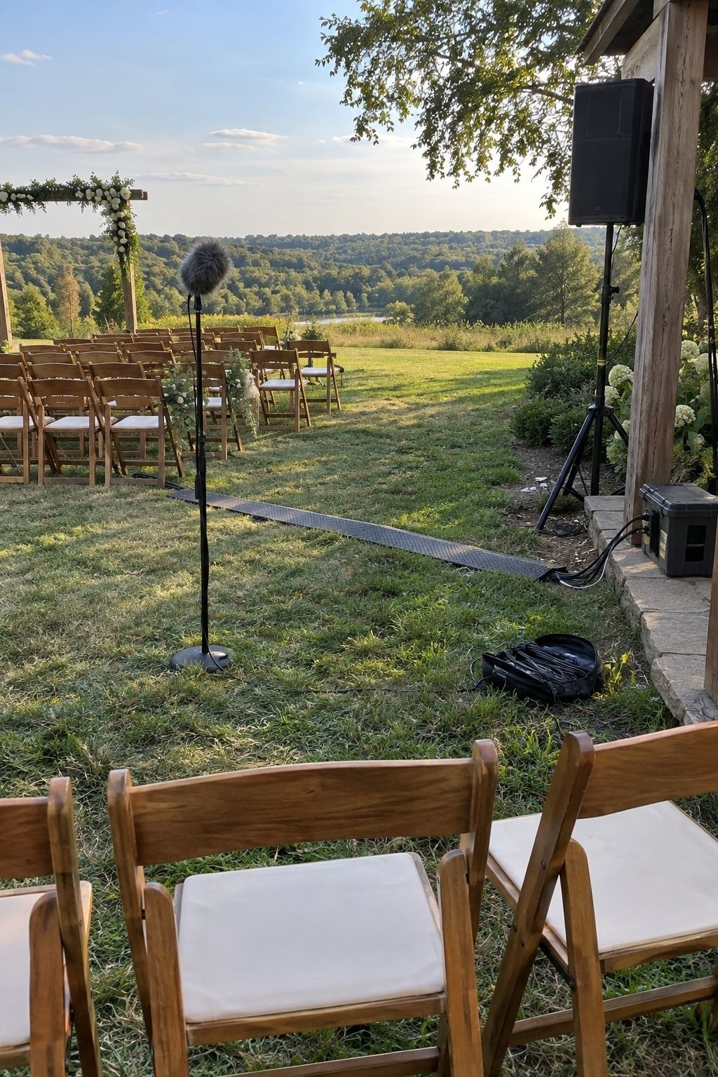 Outdoor wedding sound-check setup with windscreen microphone, speaker and protected cable route behind guest chairs