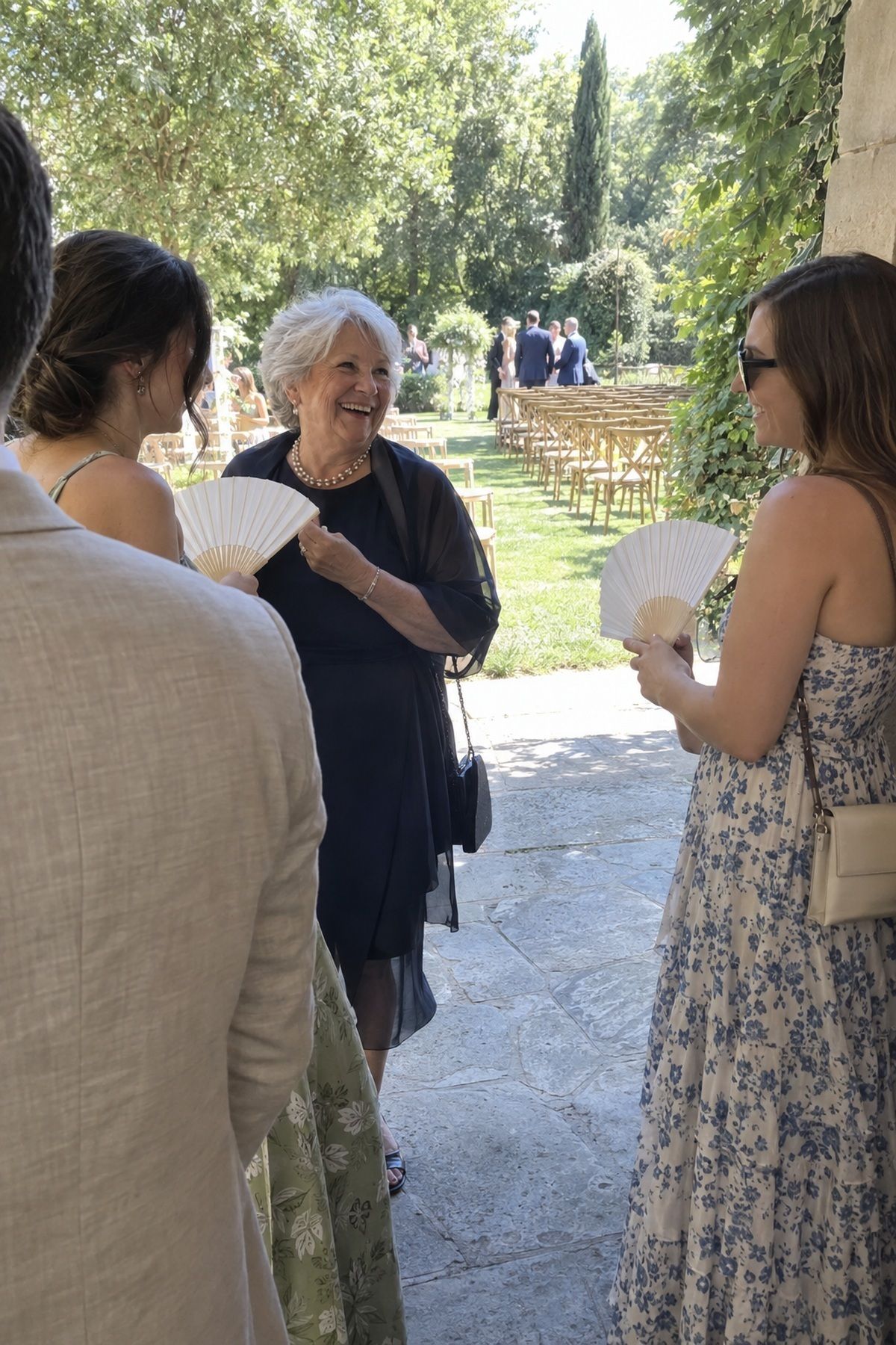 Wedding guests use paper fans while waiting in shade at an August ceremony entrance.