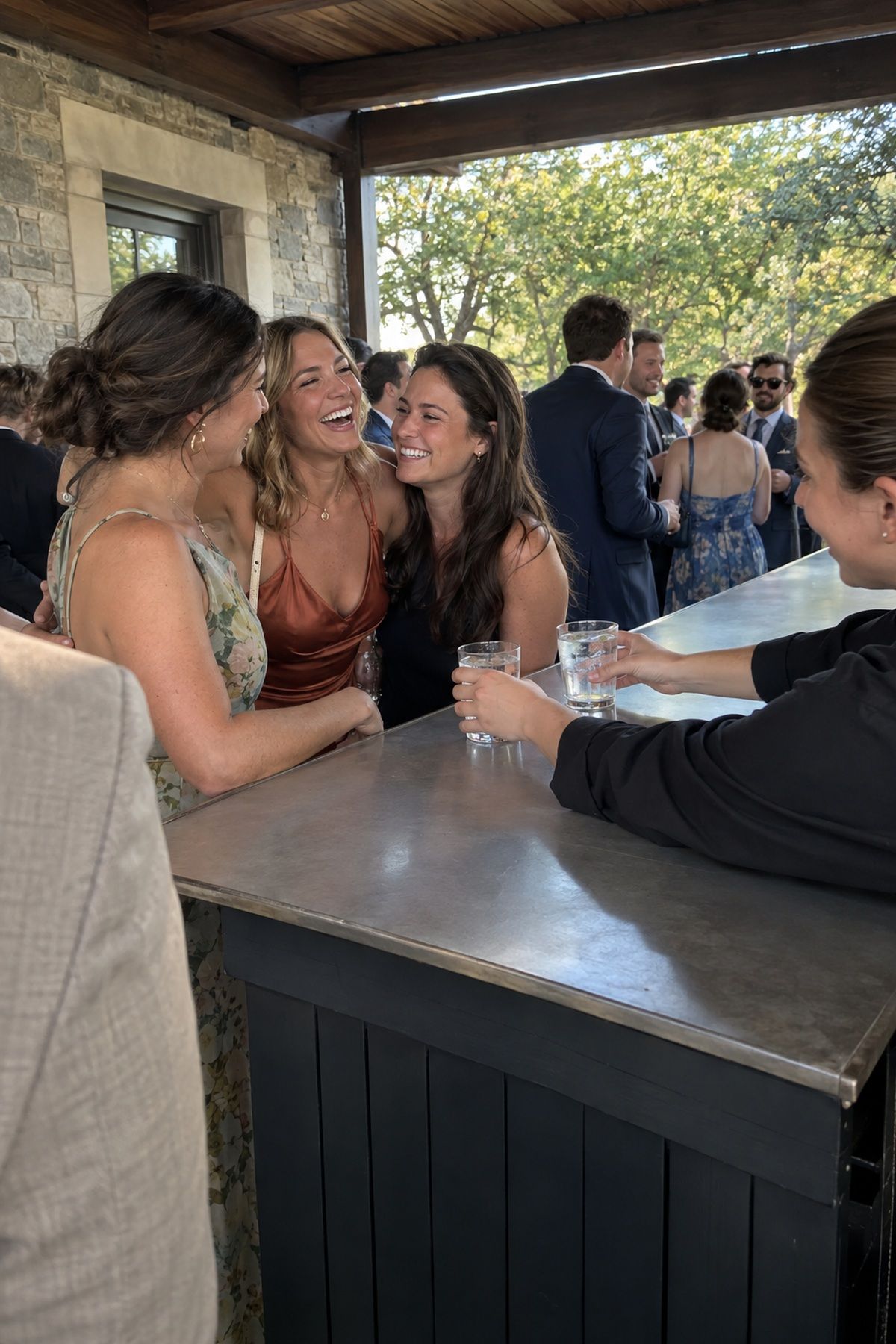 Friends laugh together beside a clean shaded bar at an August wedding.