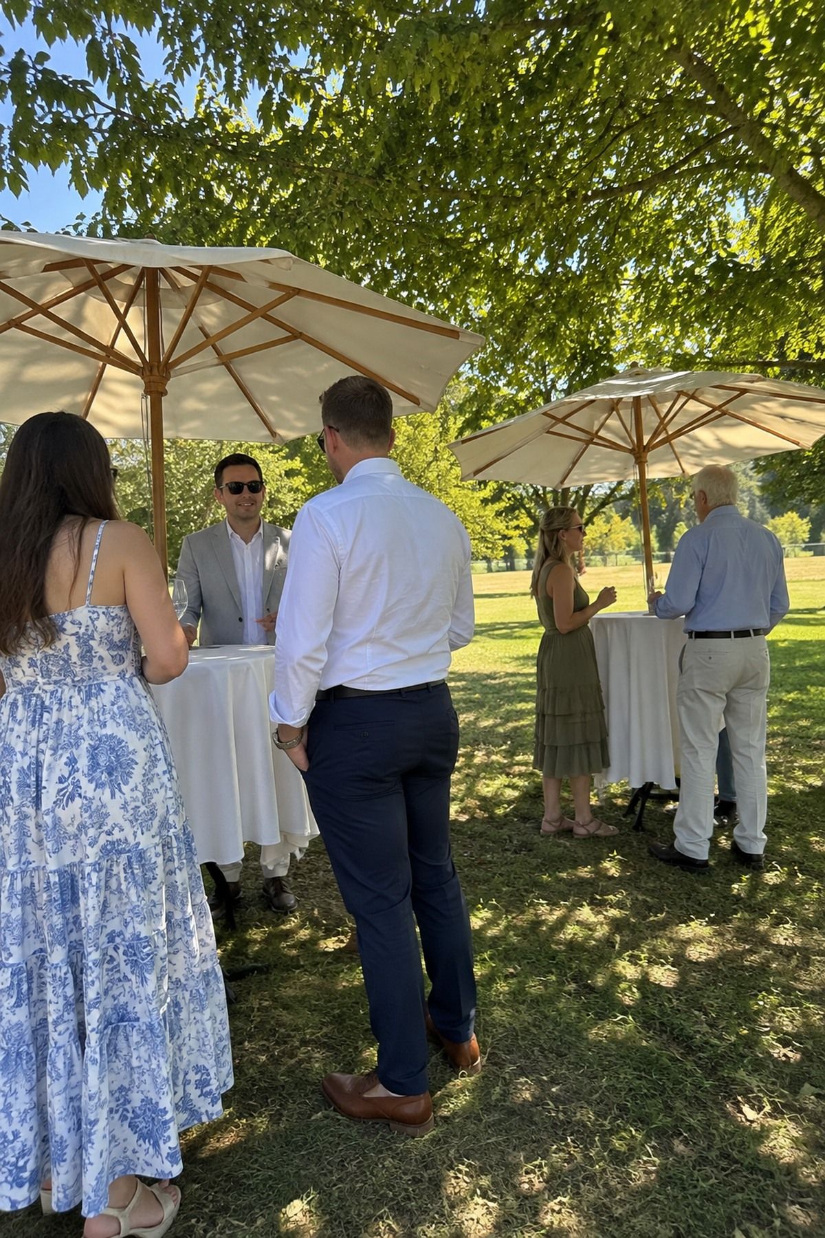 Wedding guests mingle beneath market umbrellas and mature trees during a shaded August cocktail hour.