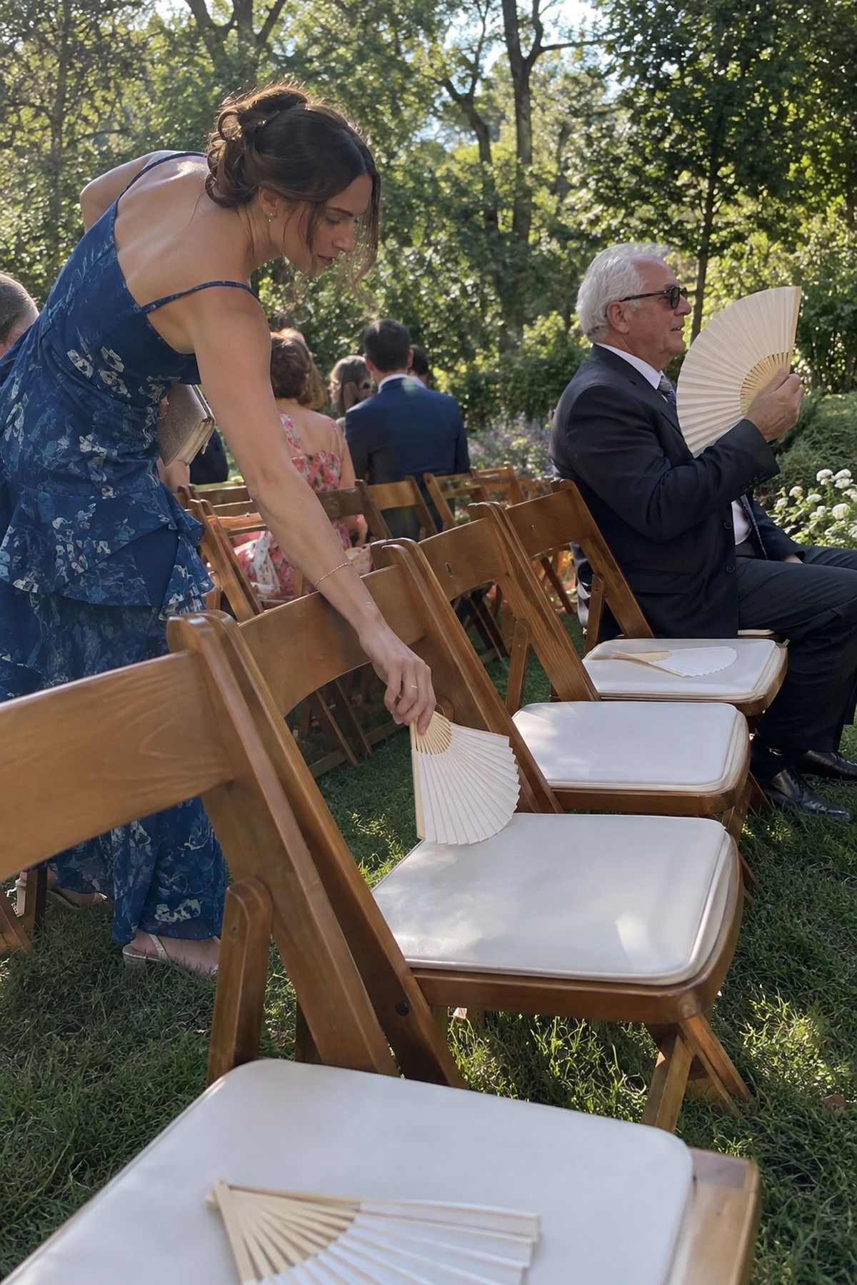 Guests pick up plain paper fans from garden ceremony seats before an August wedding.