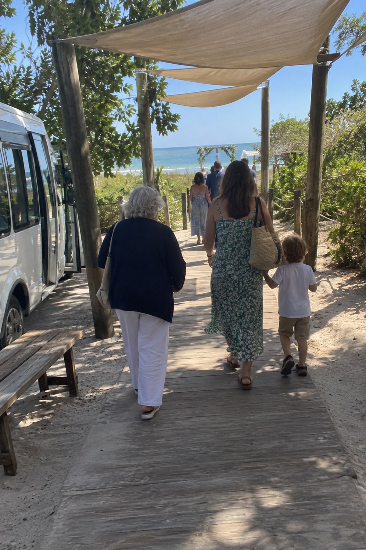 Guests walk a short shaded path from a shuttle drop-off to a beach wedding ceremony.