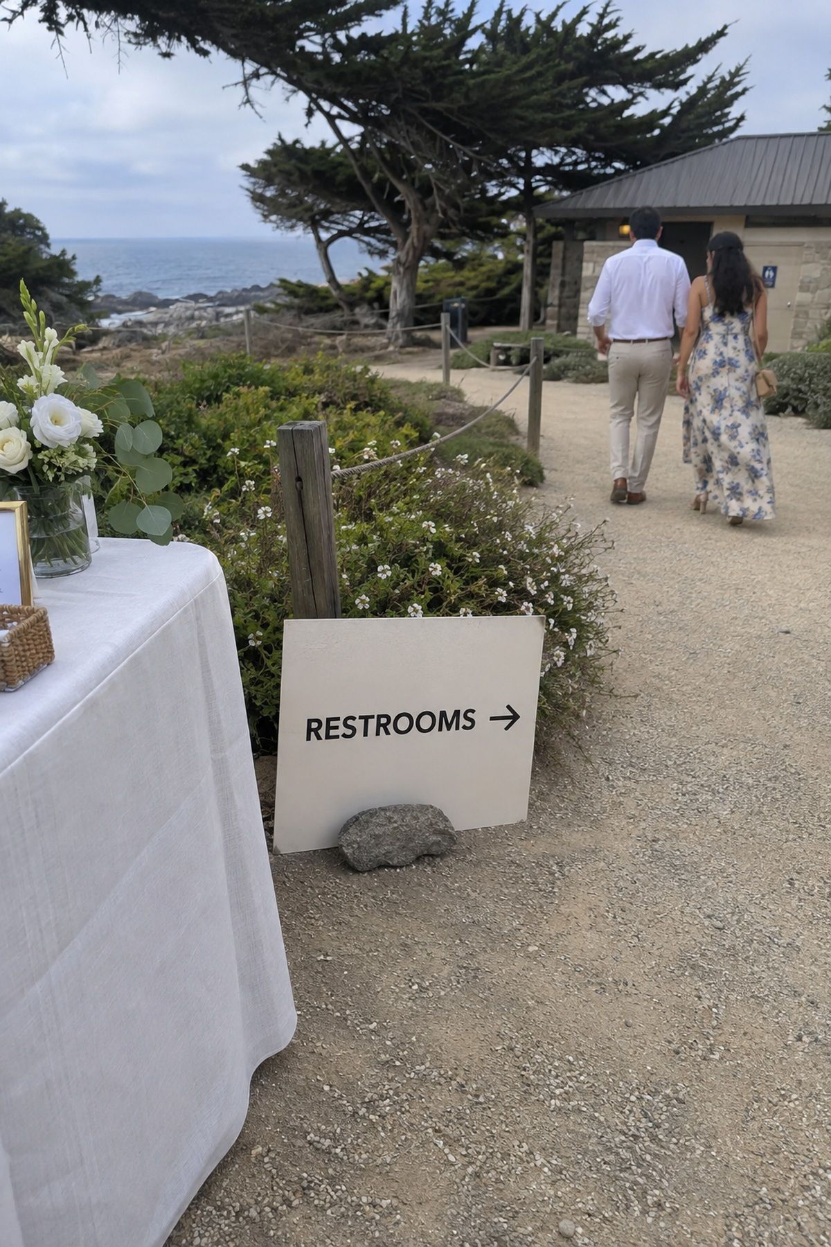A clear weighted restroom sign points beach wedding guests toward nearby facilities.