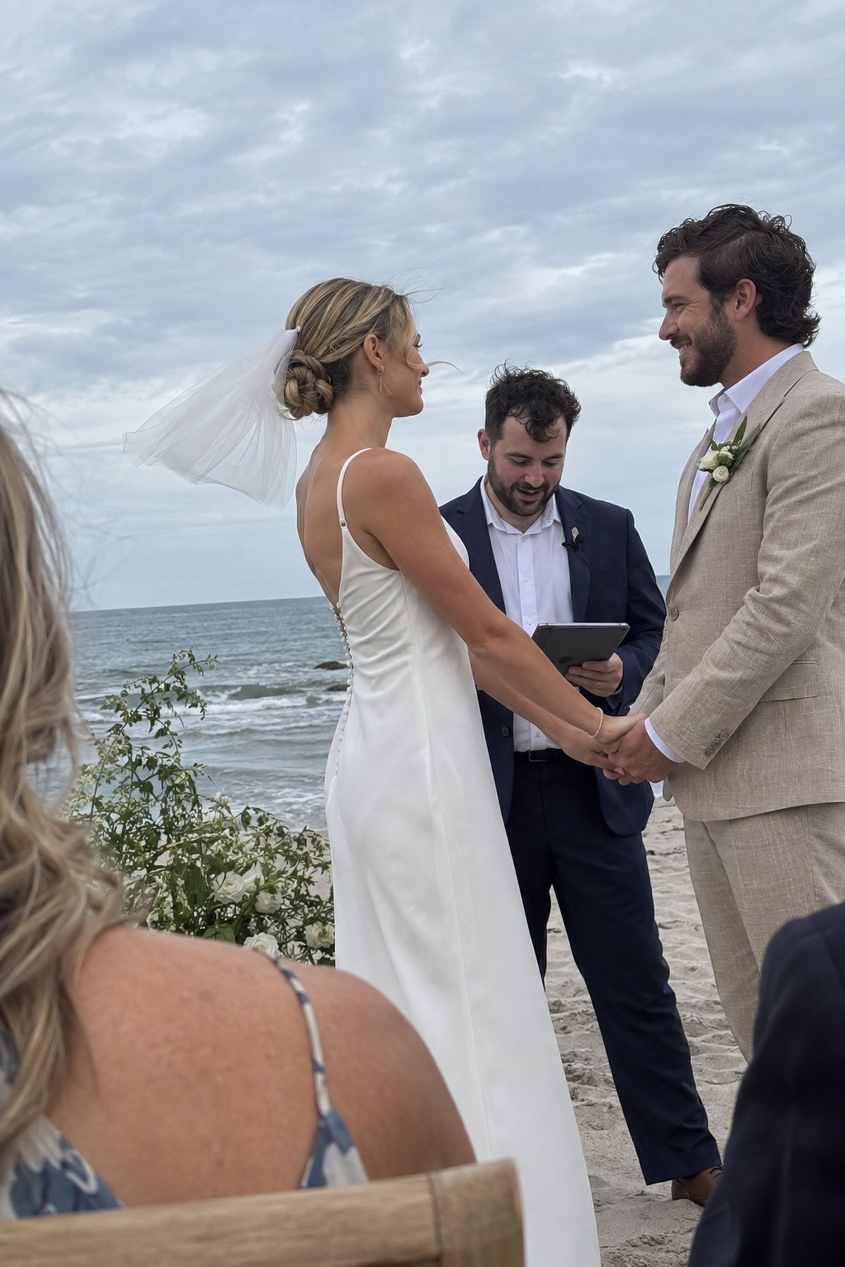 A bride wears a secure low bun and short clipped veil during a breezy beach ceremony.