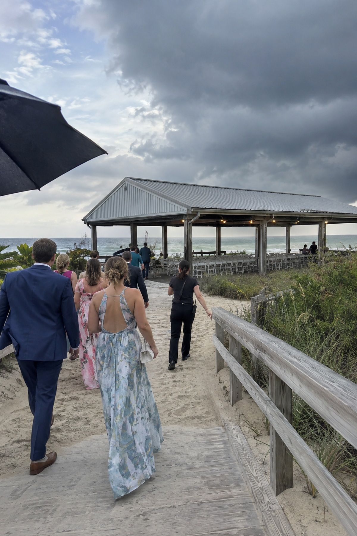 Beach wedding guests walk to a nearby covered pavilion as rain clouds gather offshore.