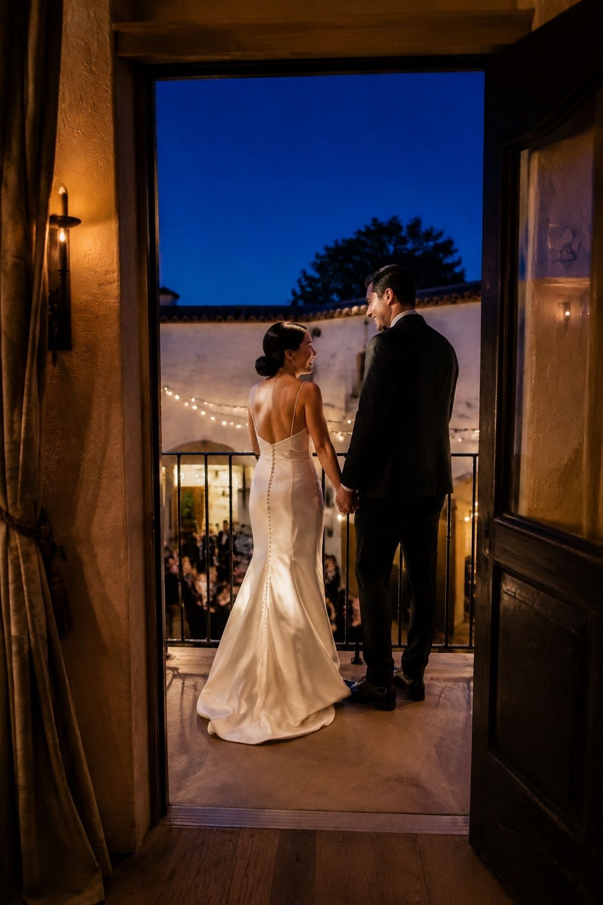 Newlyweds holding hands on a balcony above their warm-lit blue hour reception