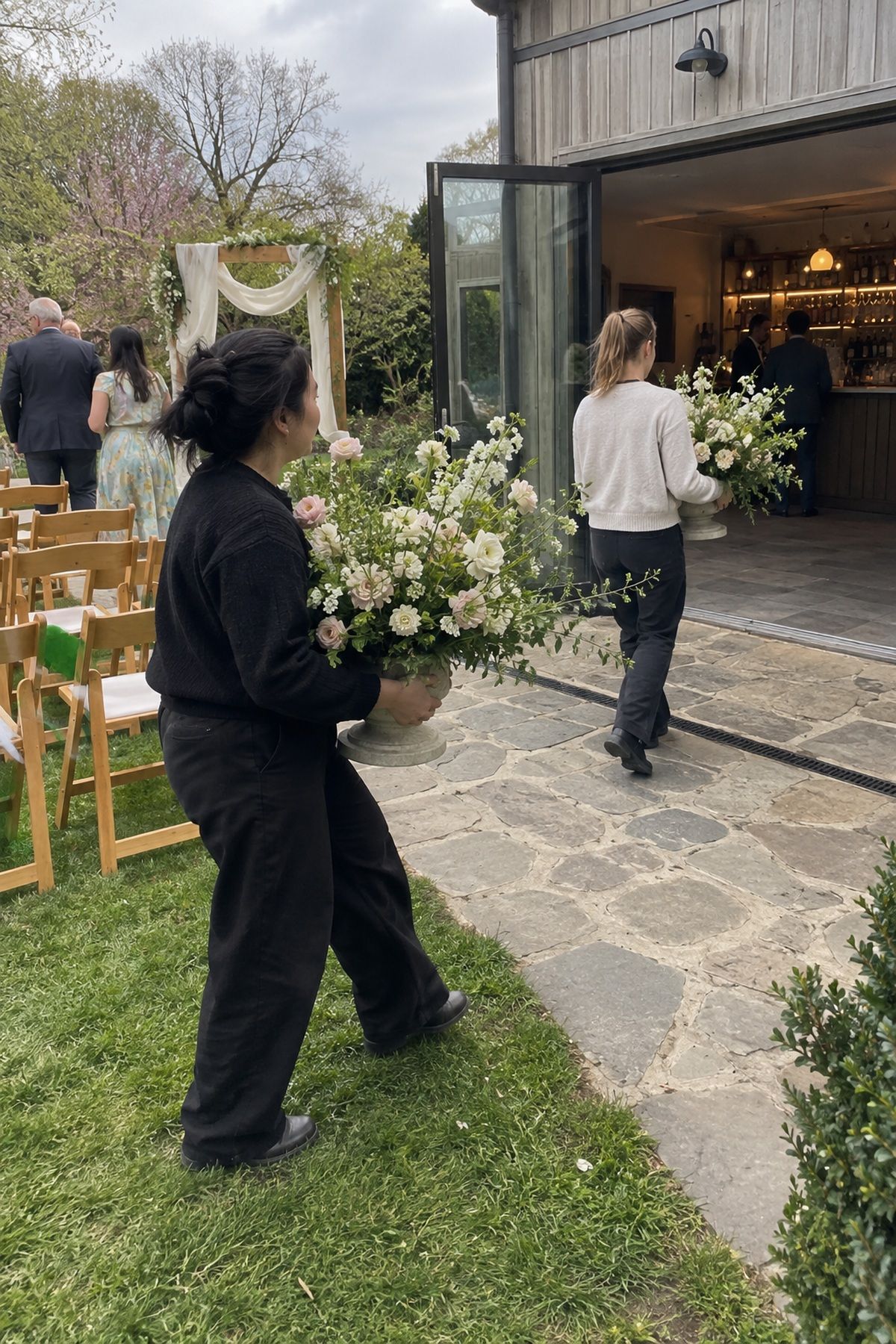 Wedding helpers move ceremony flowers toward the reception bar for a second use.