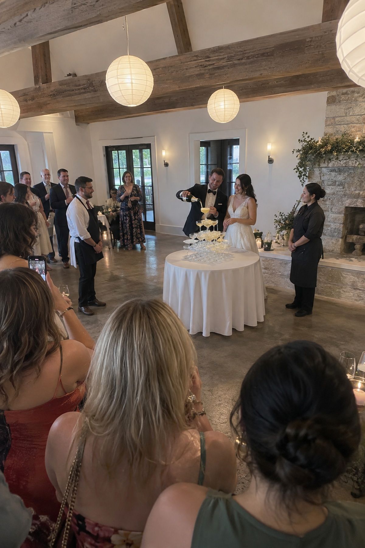 Wedding guests gather in a safe open arc around a champagne tower pour.