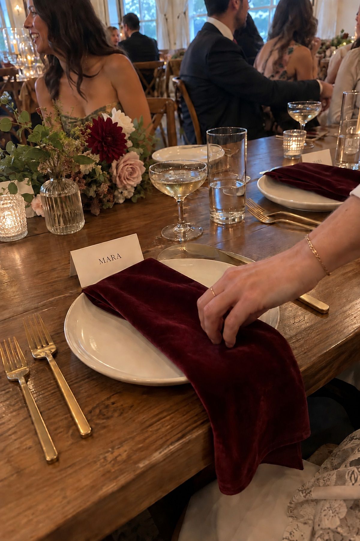 A guest adjusts a cranberry velvet napkin beside a simple winter wedding place setting.