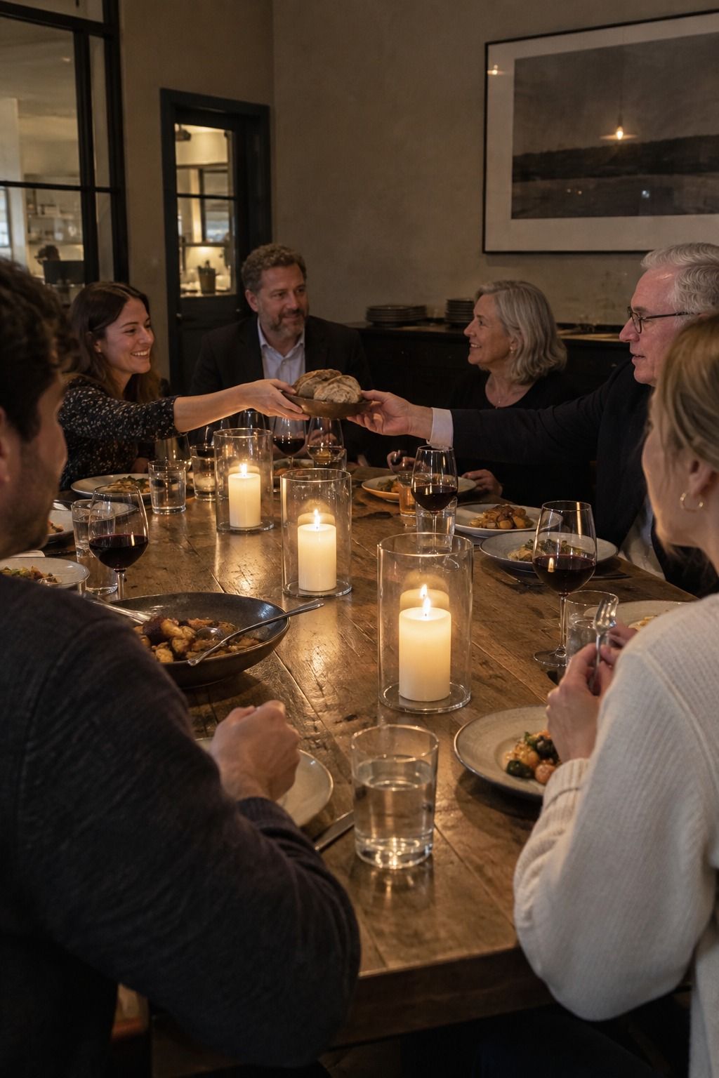 Six mixed-age guests pass bread around three low ivory candles, each flame fully enclosed by a tall stable clear hurricane sleeve.