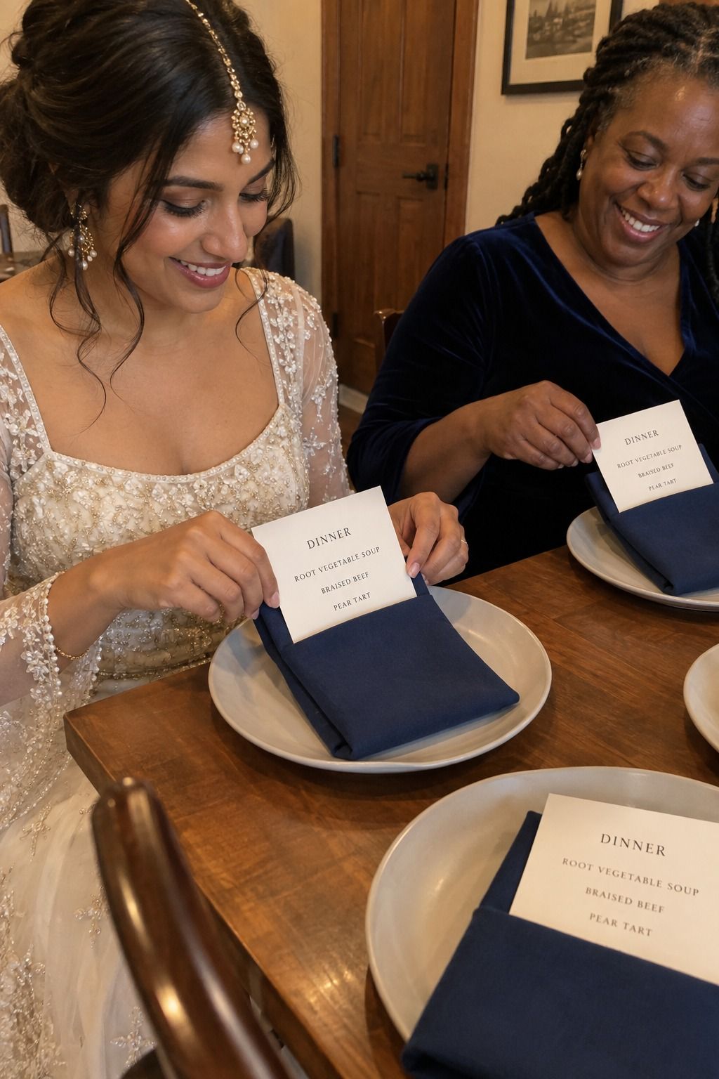 A South Asian bride and Black friend tuck readable dinner menus into deep-blue napkins; every visible card correctly lists dinner, soup, beef, and pear tart.