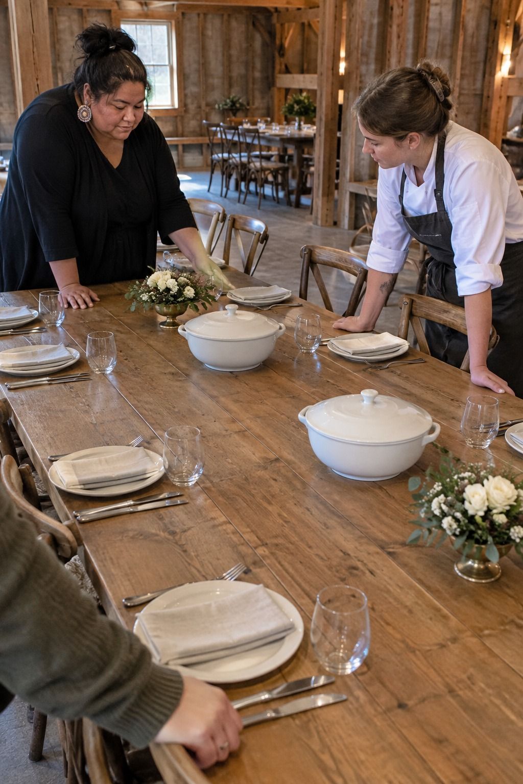 A coordinator and caterer test low flower compotes against empty covered serving tureens in wide bare gaps on a long barn table.