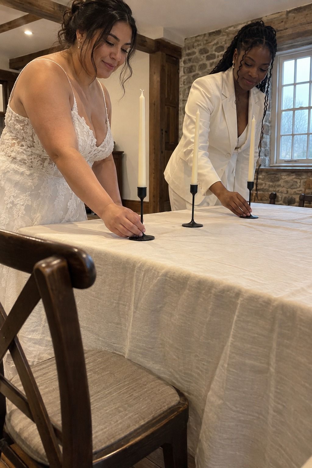 Two brides space three slim matte-black holders with unlit ivory tapers across softly wrinkled cream linen.