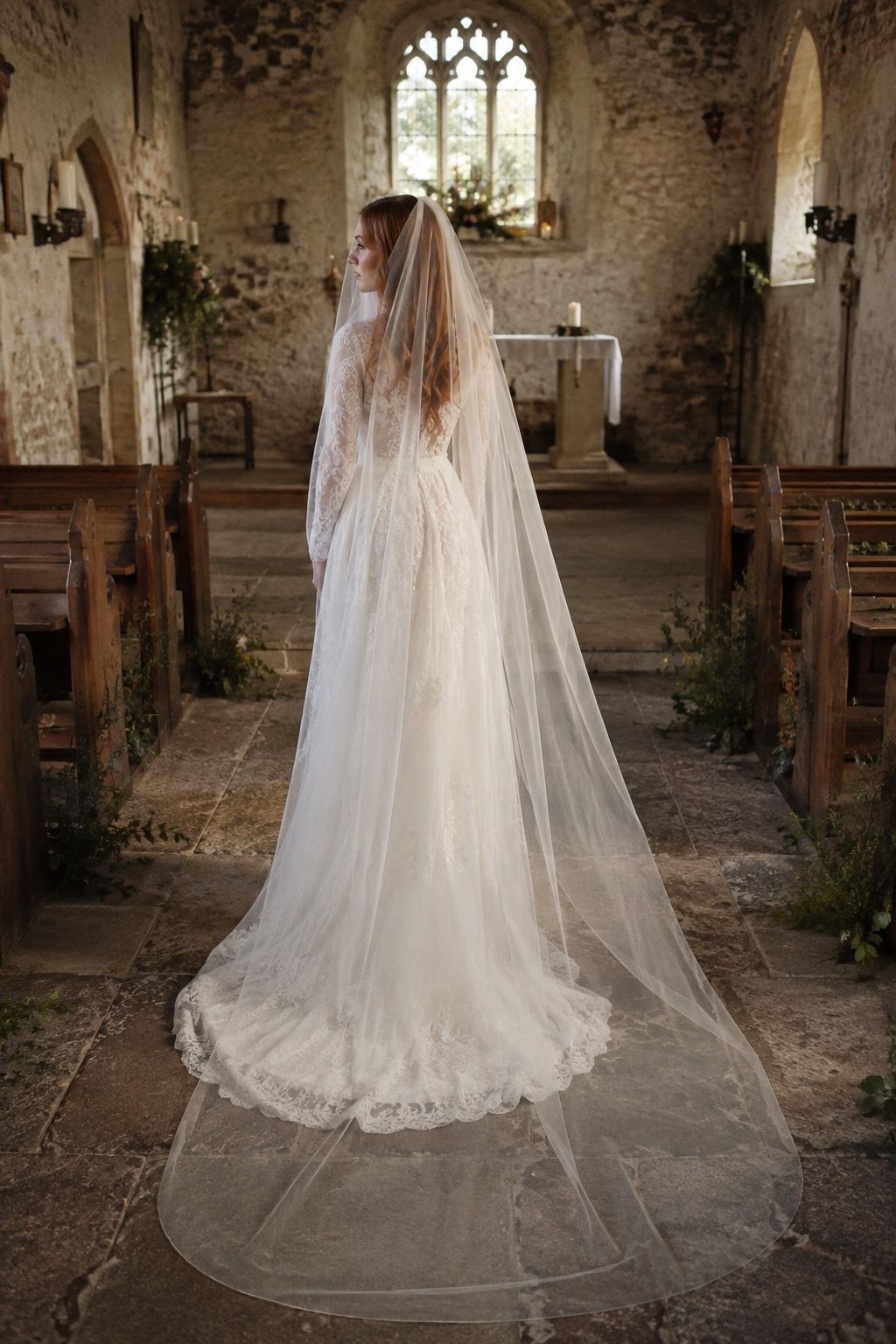 Wide rear three-quarter full-length red-haired bride in small stone chapel aisle, flat cathedral drop veil extending two meters along floor, sheer front blusher covering profile