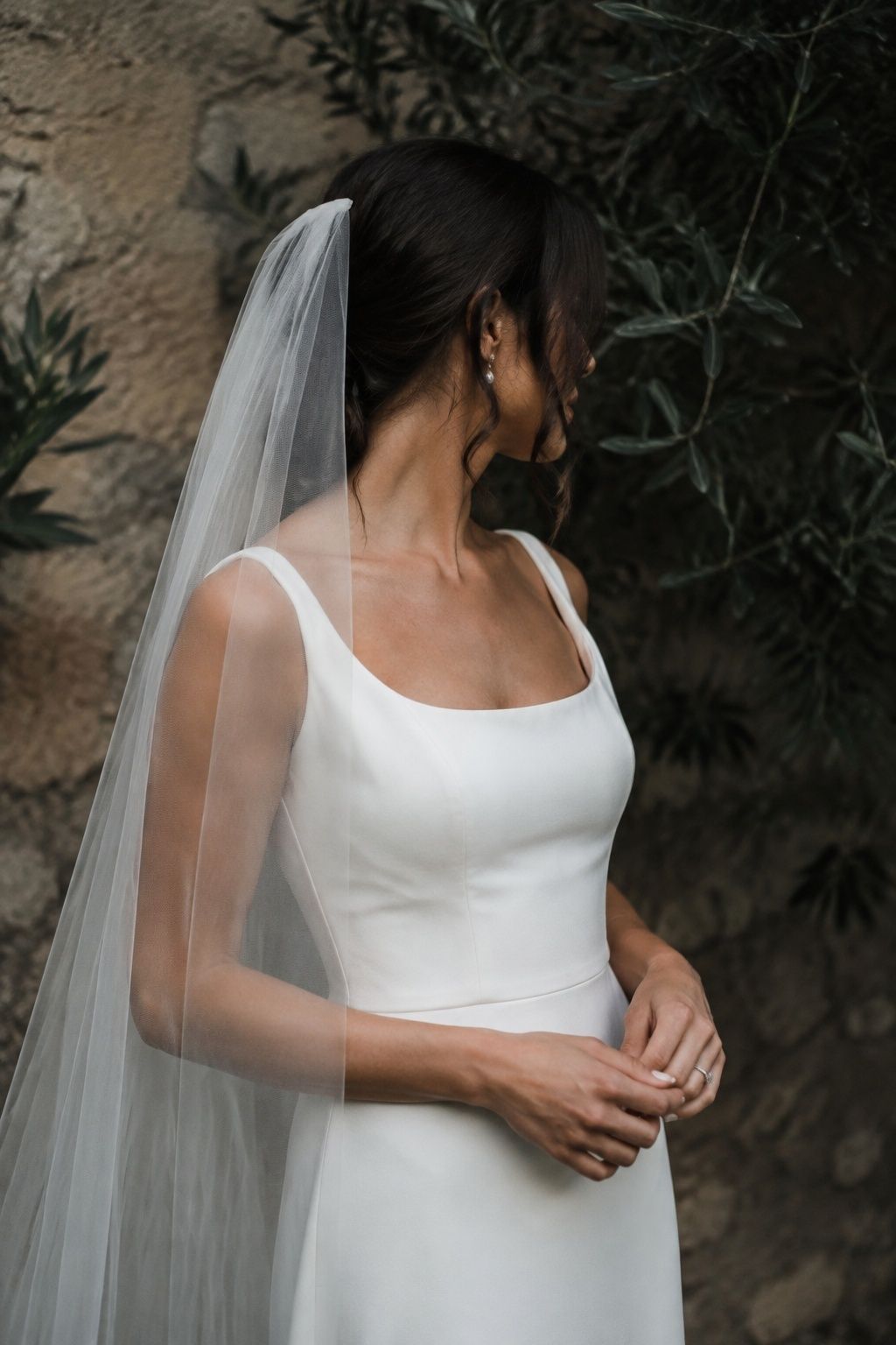 Close side waist-up bride against olive garden wall, cleanly cut raw tulle edge visible at shoulder against dark foliage, flat crown and modern crepe gown