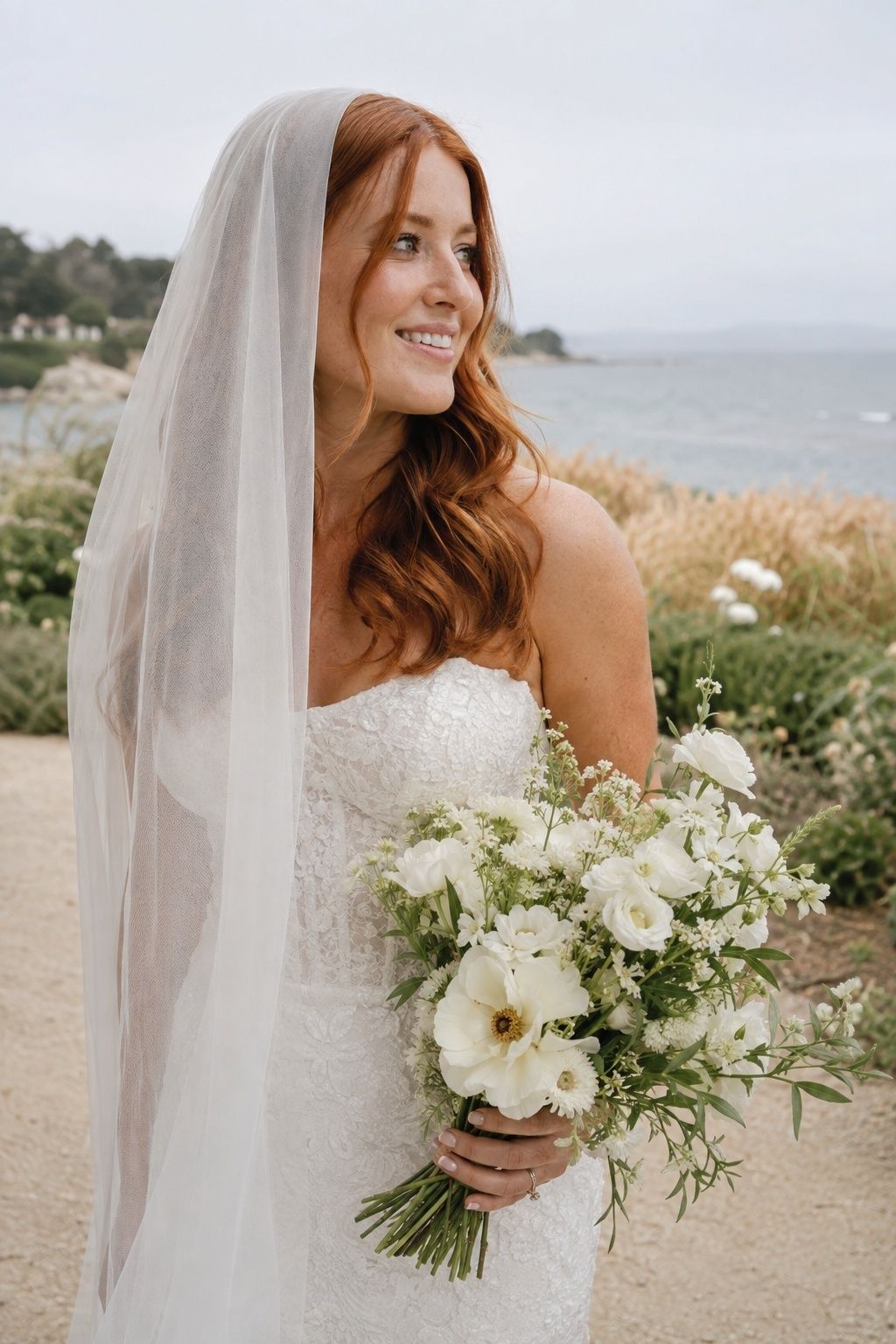 Waist-up side portrait of auburn bride with loose waves beneath ungathered sheer drop veil in breezy seaside garden, actual face clearly visible