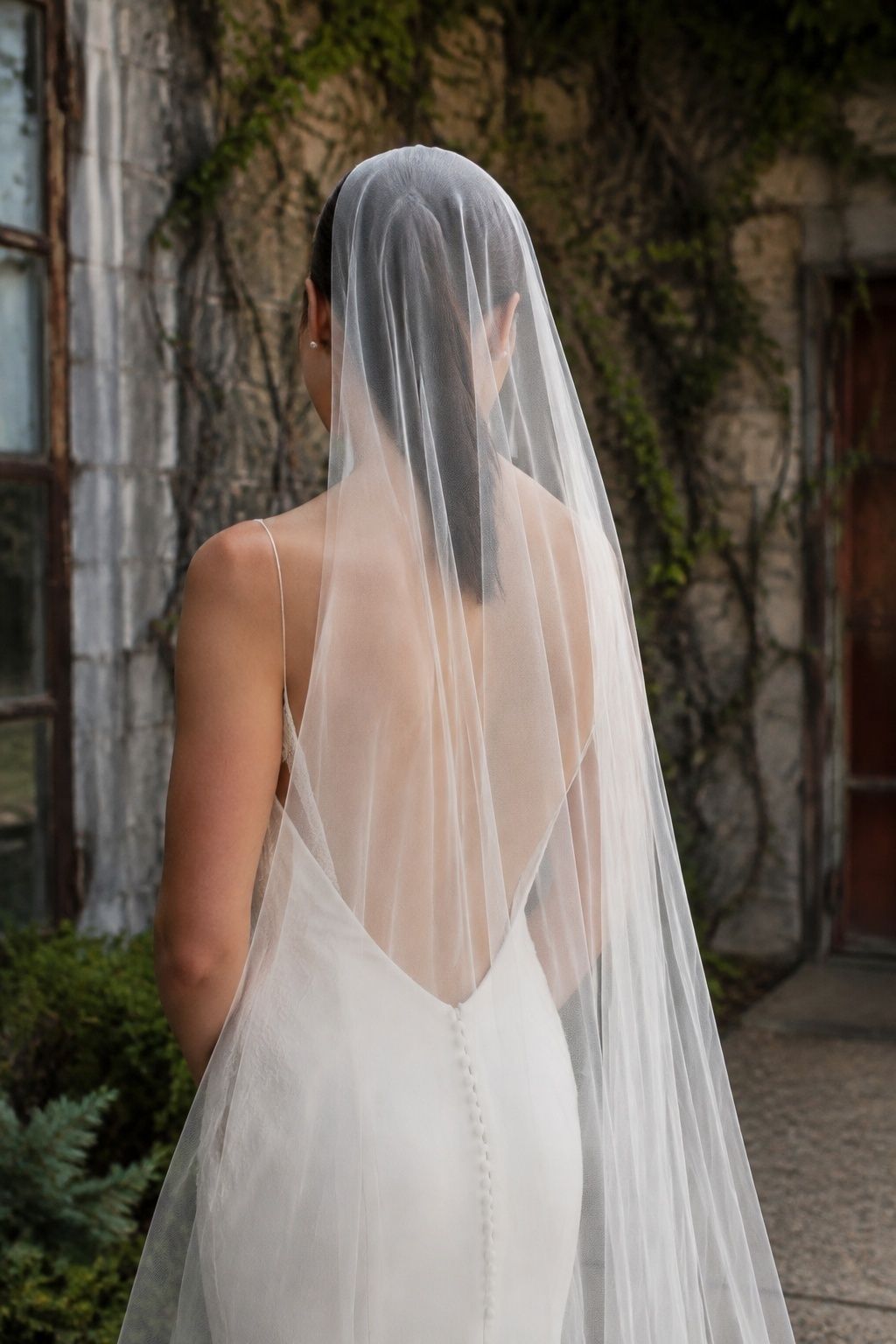 Rear waist-up bride with ivory open-back gown and small covered buttons, exceptionally sheer ungathered veil allows low back design to remain clearly visible