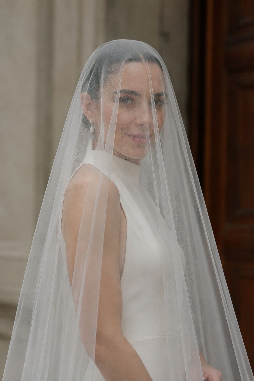 Front three-quarter half-length bride with high-neck ivory crepe gown and delicate earrings, flat sheer face blusher softly framing shoulders without volume
