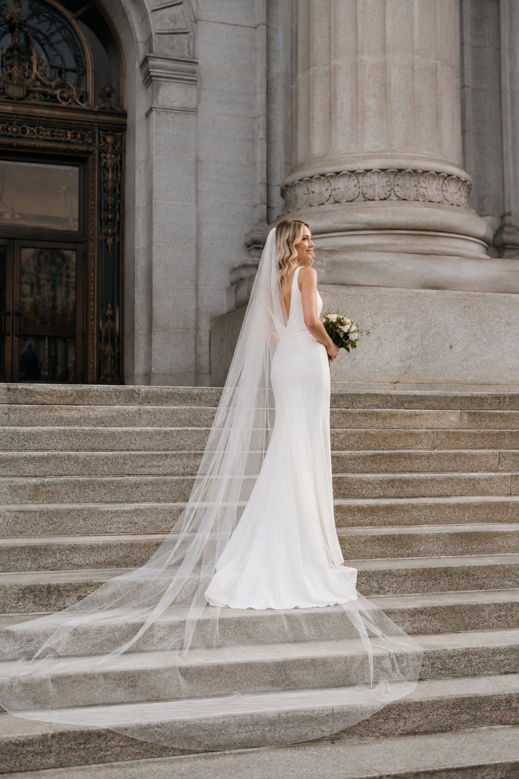 Full-length bride on city hall stone steps, narrow minimalist column gown and chapel-length plain drop veil, straight dress shape contrasted with airy trailing tulle