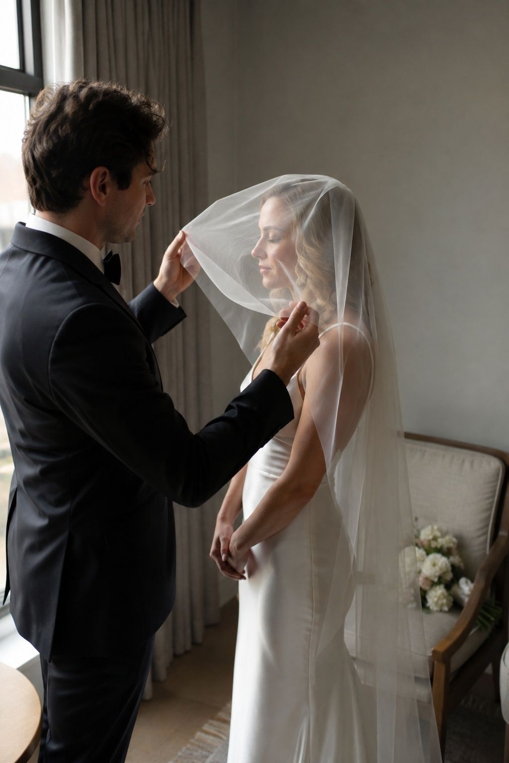Intimate side-view rehearsal of partner gently lifting front veil layer away from brides rendered face, two natural hands holding outer edges, veil securely flat at crown, bouquet rests on nearby chair