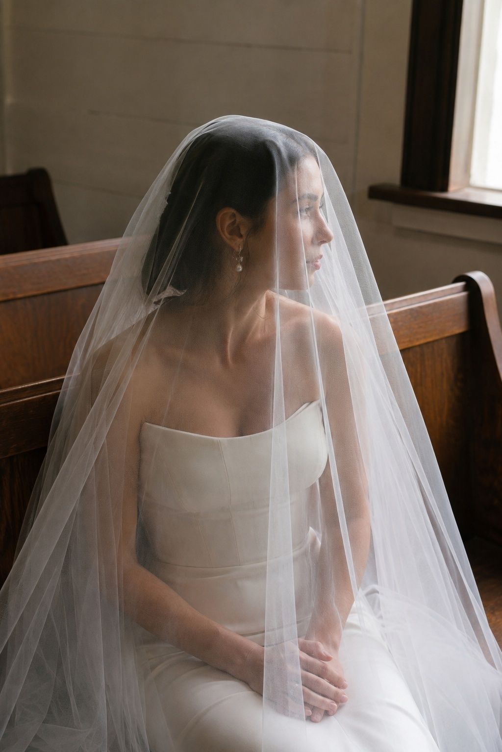 Seated waist-up bride alone in small wooden chapel pew, sheer drop veil over face, soft side window light, natural fully rendered face and relaxed hands in lap