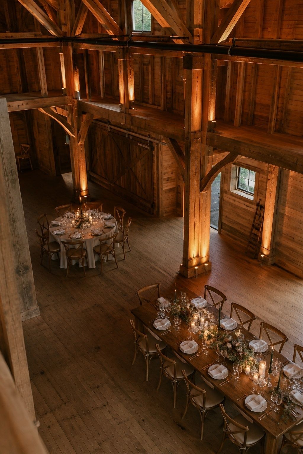 Wide upward oblique view of a modest timber barn wedding reception with subtle warm amber uplights at beam bases washing natural wood