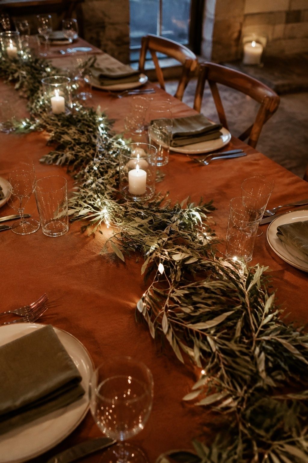 High oblique close photo of fine warm fairy-light wire woven sparingly through a low olive greenery runner on a wedding table
