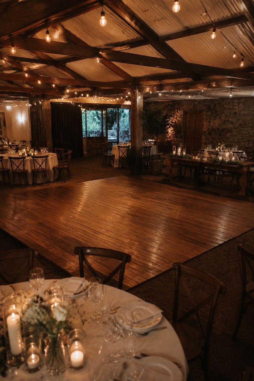 Wide slightly elevated view of a small empty wooden wedding dance floor surrounded by a gentle warm-white light wash and soft overhead bulbs