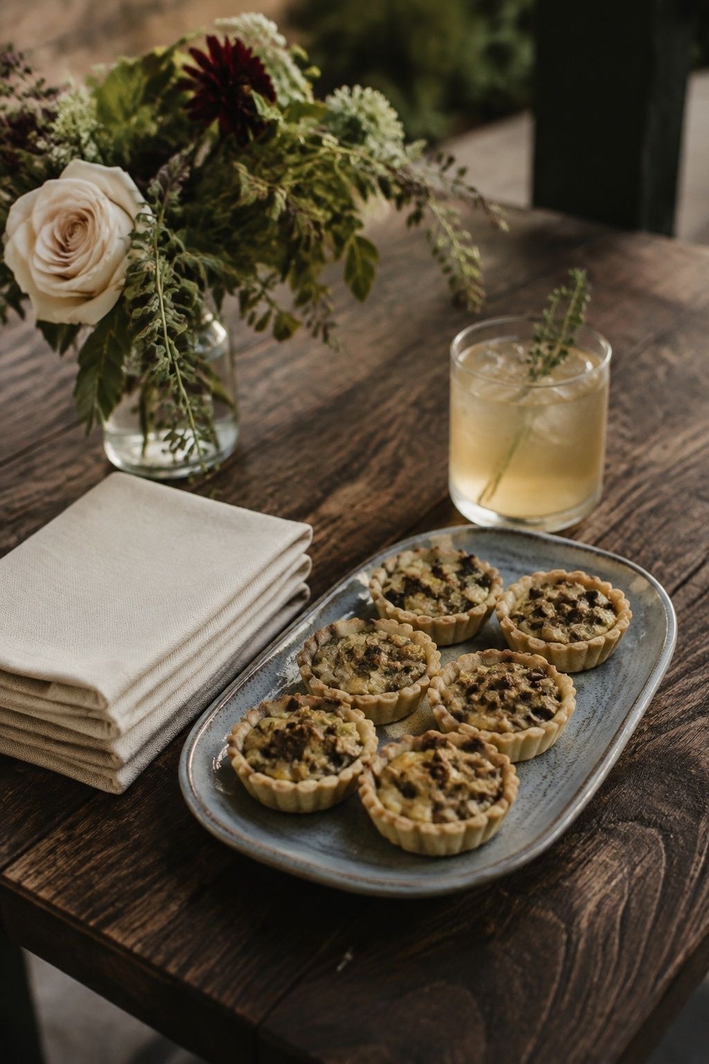 Six tiny leek and mushroom savory tartlets with flaky pastry on a blue-gray ceramic platter, ivory napkins beside a welcome drink at a small fall wedding, intimate close side view