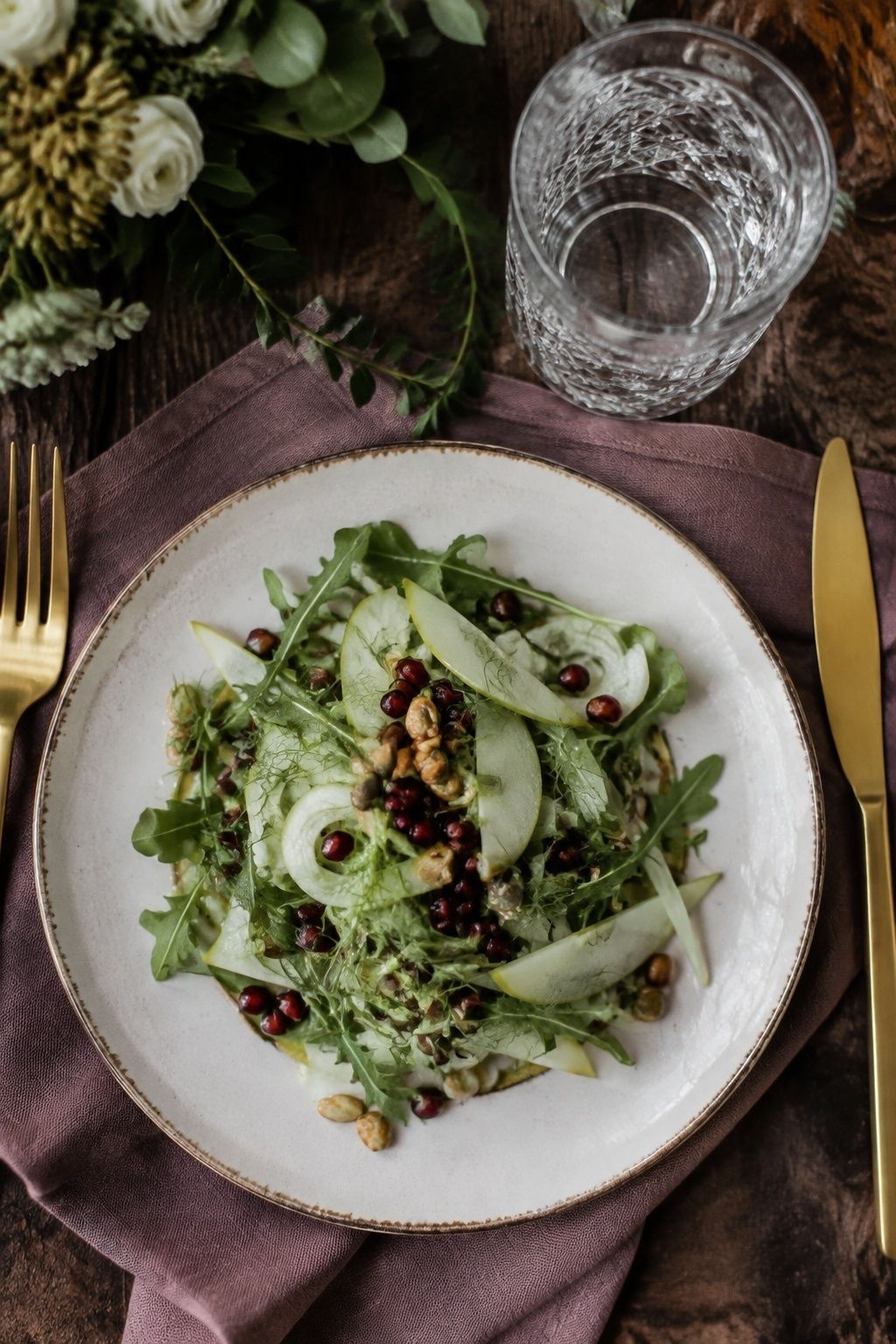 Fresh arugula pear and shaved fennel salad with pomegranate and toasted seeds on an ivory starter plate, bright cider vinaigrette, plum linen napkin and wedding water glass, overhead detail