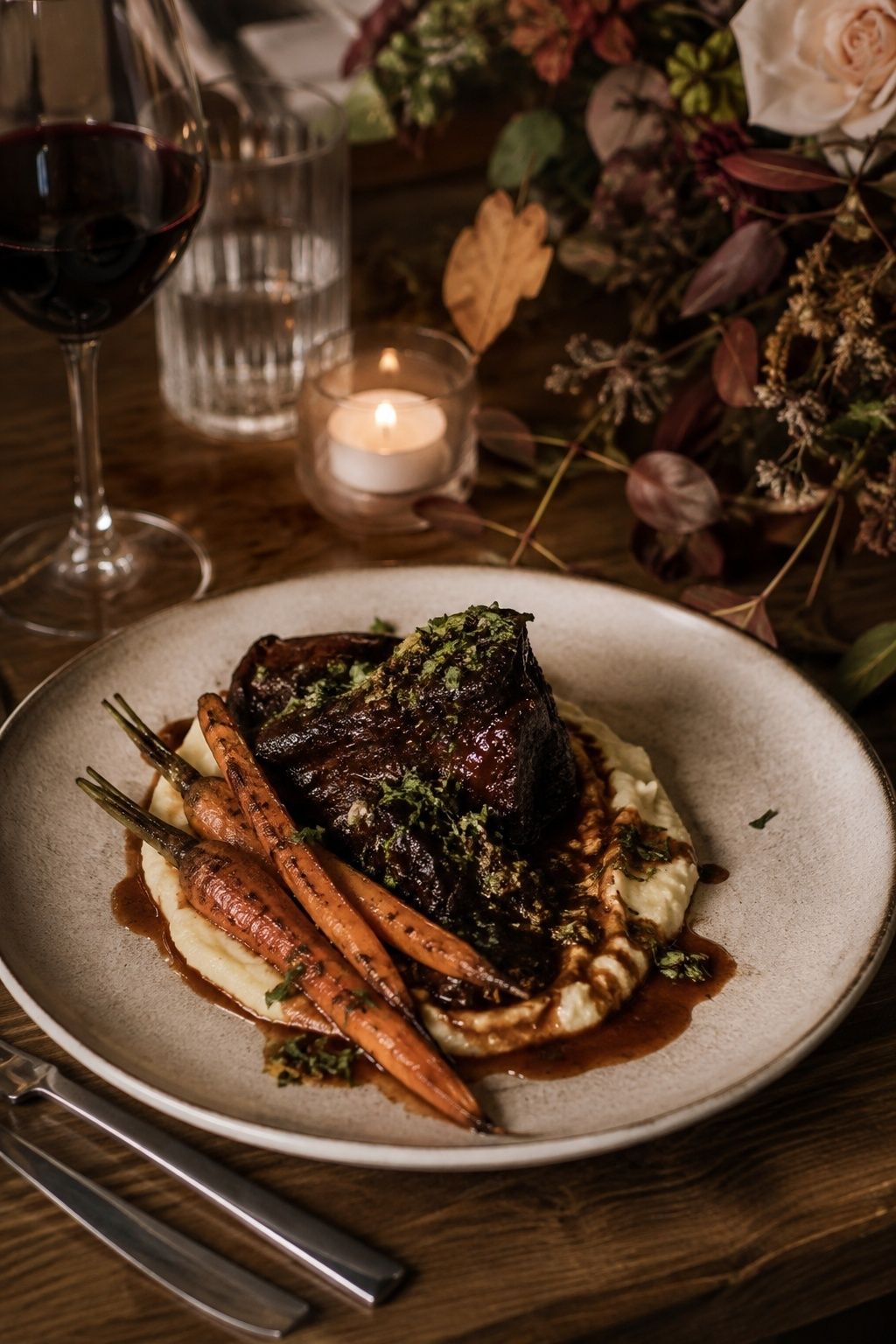 Braised short rib on creamy polenta with roasted carrots and bright green gremolata, restrained red-wine jus on a pale stoneware dinner plate at an intimate autumn wedding, low close view