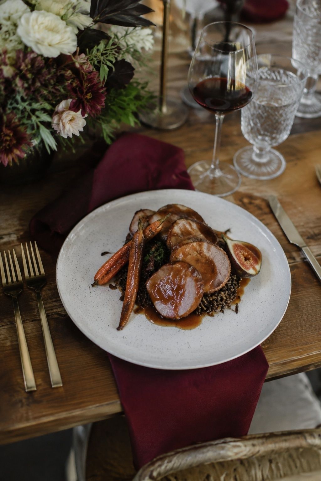 Sliced pork tenderloin with restrained glossy fig glaze, halved fig, wild rice and roasted carrots on a wedding dinner plate, burgundy napkin and clear stemware, high diagonal view