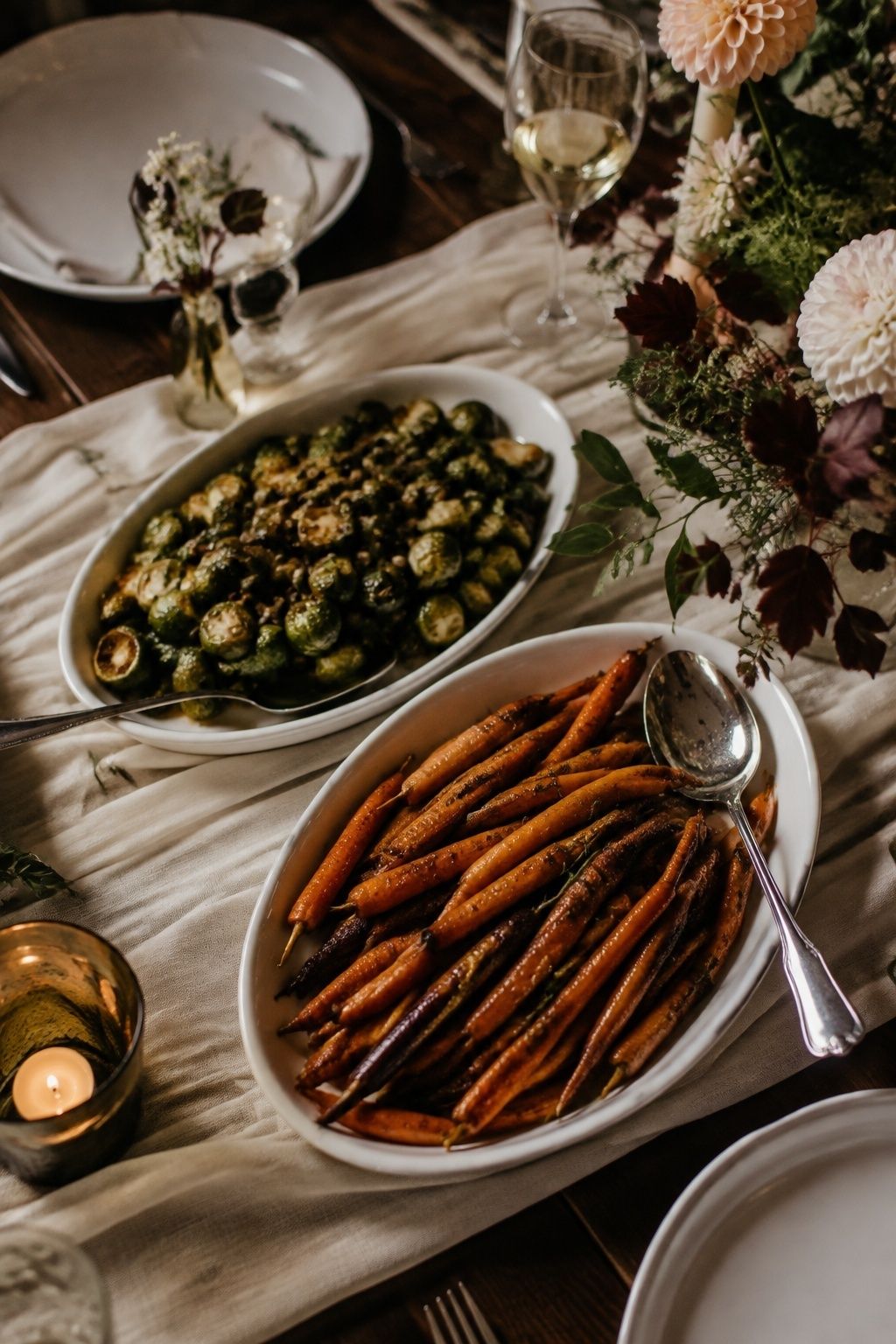 Two small family-style serving platters of roasted Brussels sprouts with toasted seeds and maple thyme carrots on a intimate wedding dinner table, practical serving spoons, colorful autumn vegetables dominant, oblique view