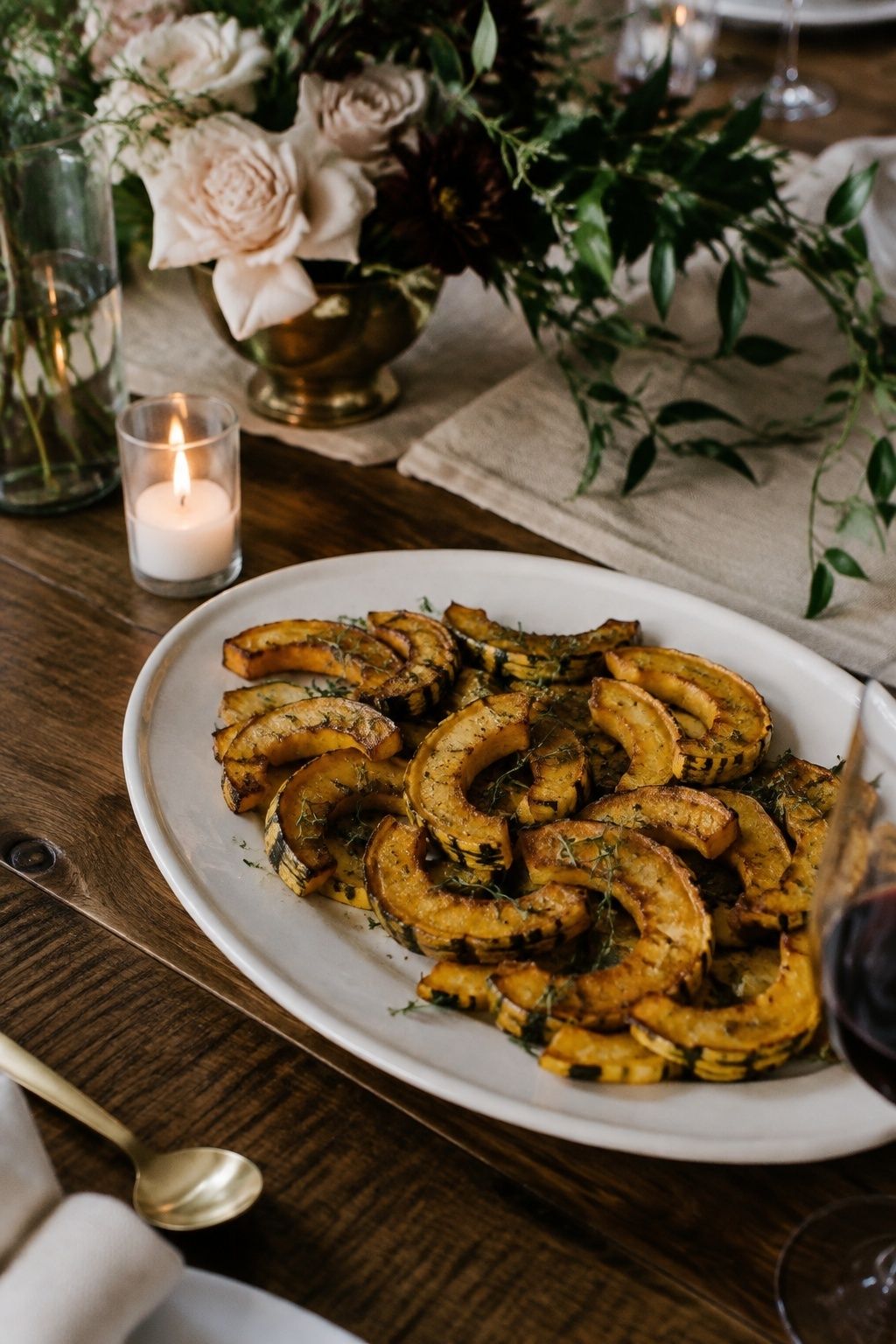 Roasted delicata squash crescent slices with thyme as the single seasonal starch on a modest white oval serving platter at an intimate wedding table, visible golden cut surfaces and green-striped rind, close tabletop view