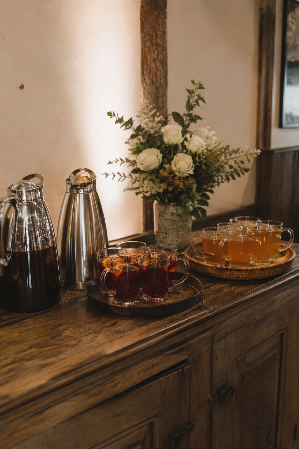 Small reception bar with two glasses of ruby mulled wine, orange peel and star anise, zero-proof amber cider in clear handled mugs on a separate tray, stainless insulated servers, warm plausible ambient light, wide straight-on view