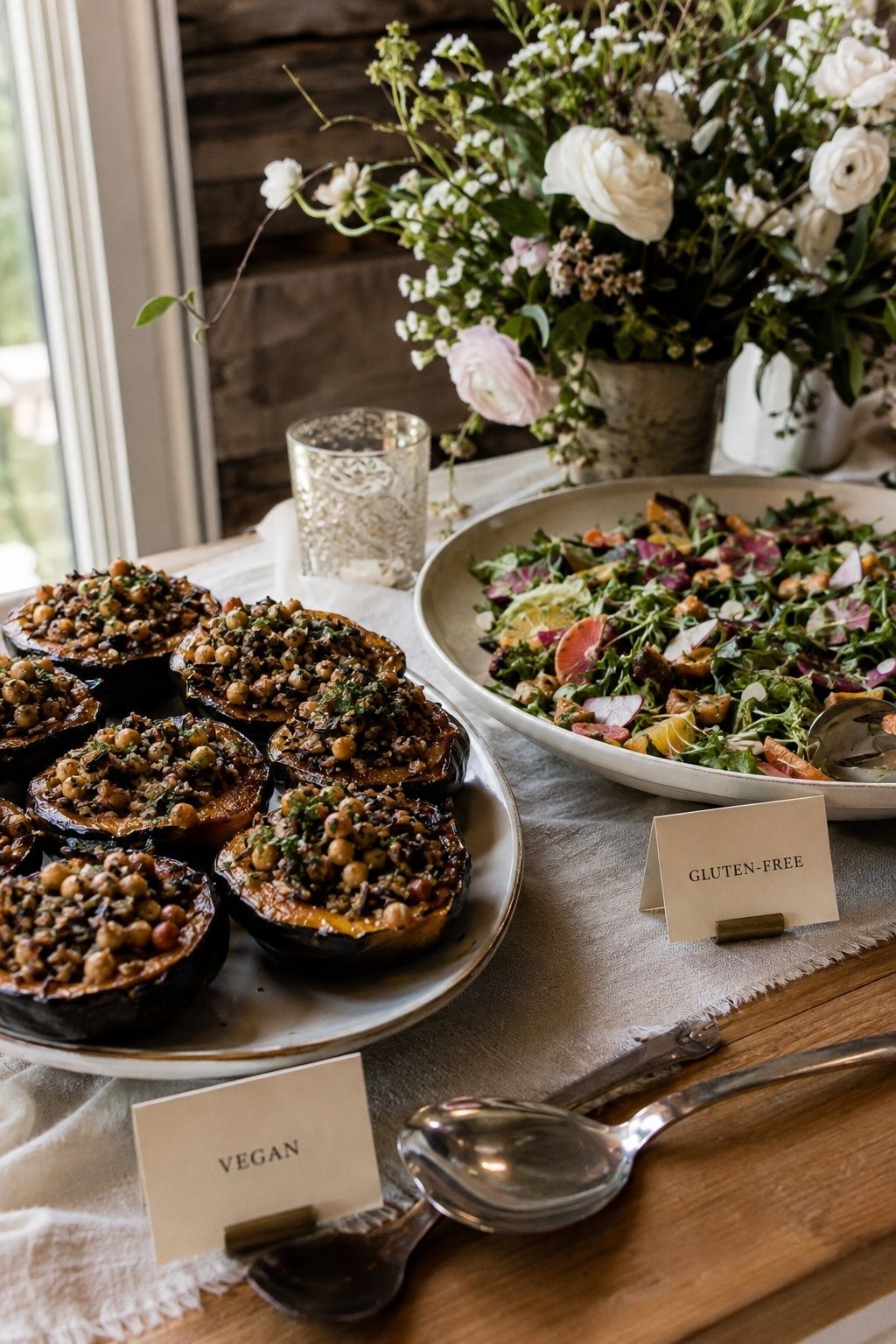 Well-organized intimate wedding buffet detail with roasted stuffed squash and bright salad, separate serving utensils for each dish, two small legible cream food cards reading exactly VEGAN and GLUTEN-FREE, dishes dominant and labels supportive, bright natural side light
