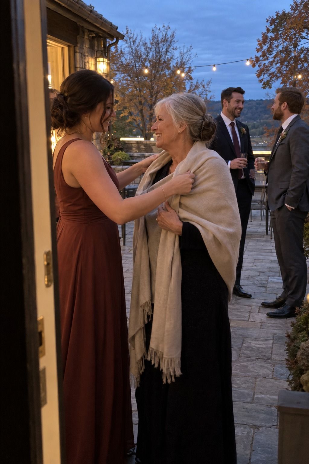 A bridesmaid helps an older wedding guest wrap up in a pashmina at dusk.