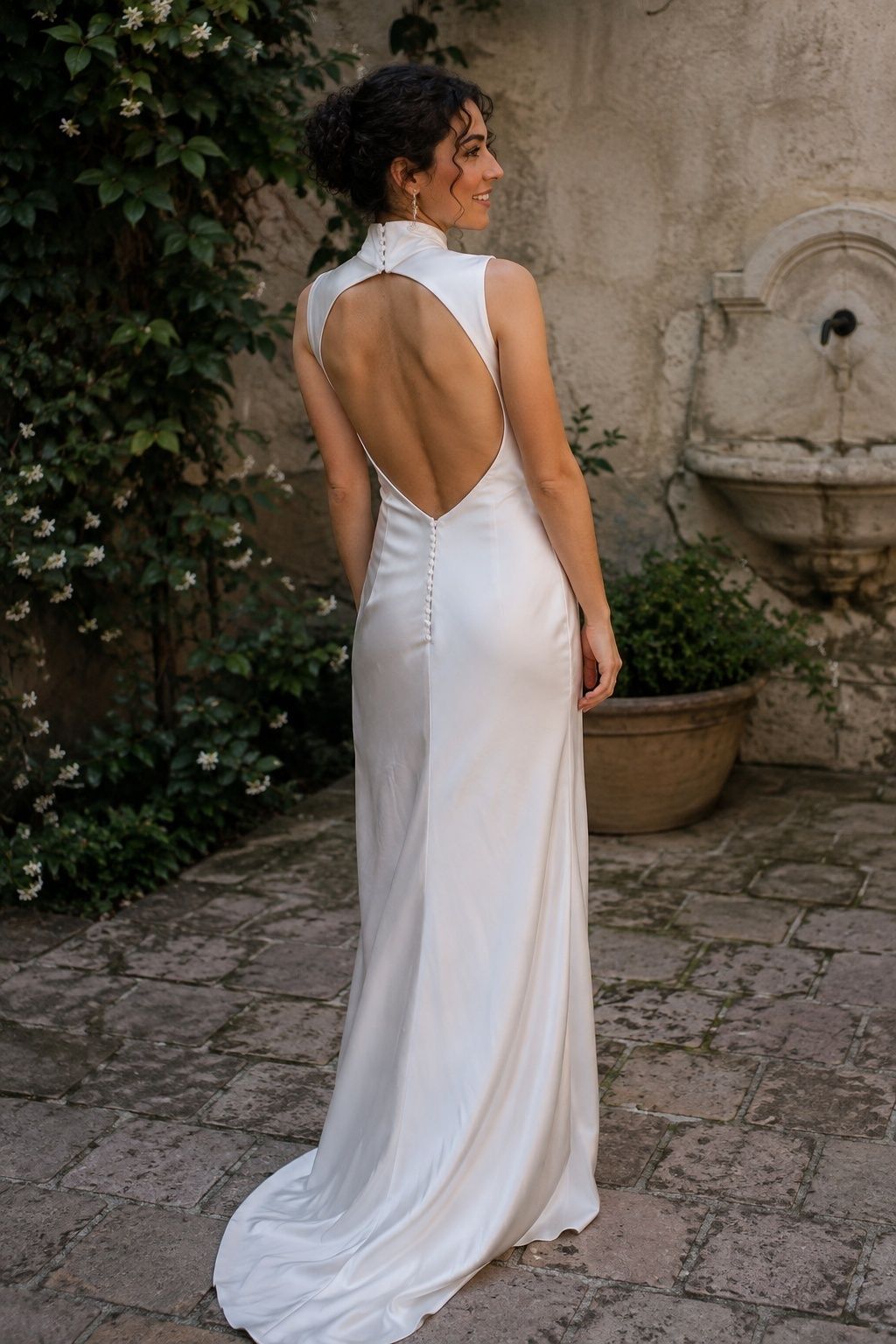 Rear three-quarter full-length adult bride with dark curly updo in an ivory gown with a high collar fastened behind neck and deep oval open back