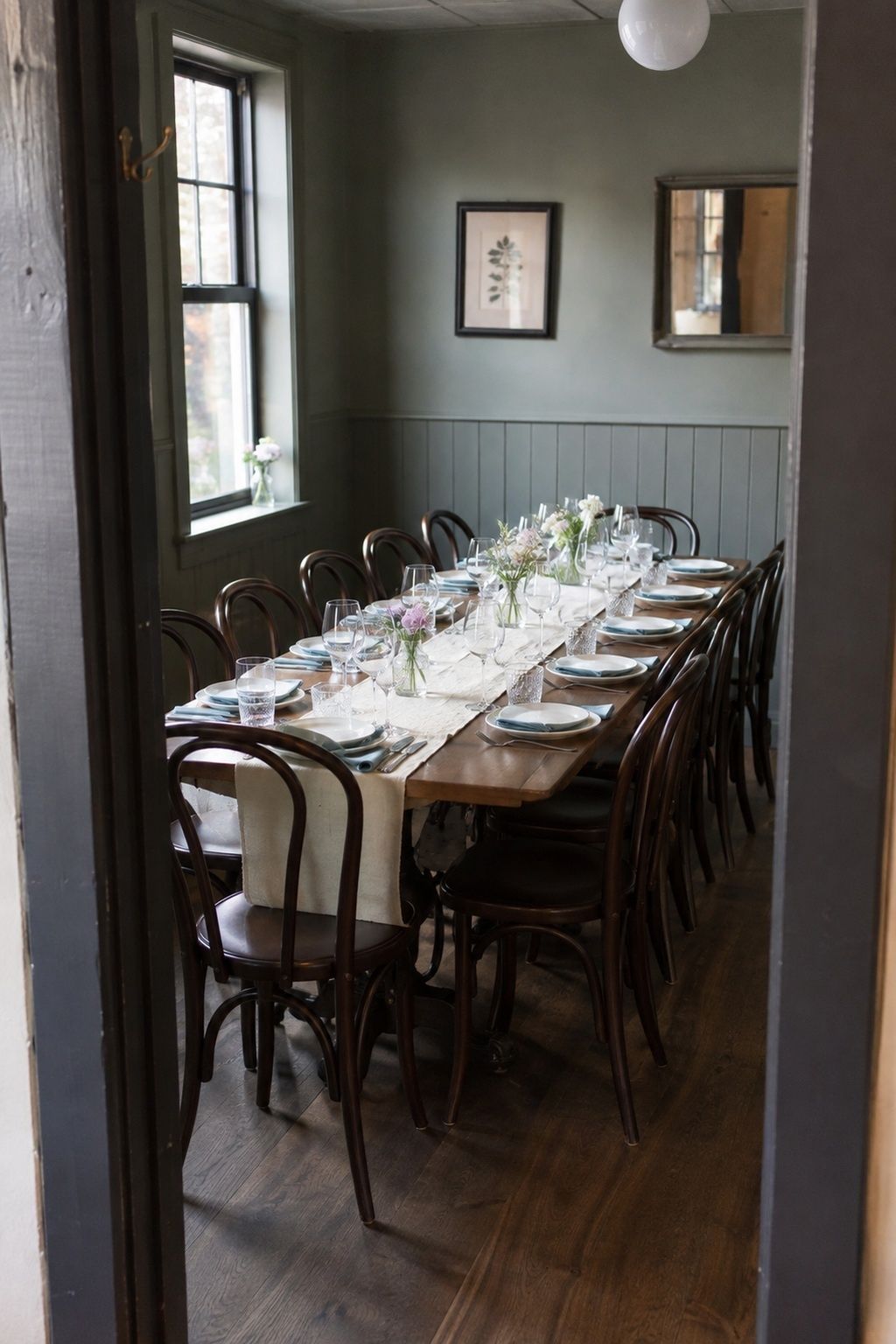 Small gray-green private dining room with one long wedding table, bentwood chairs, white place settings and small flower arrangements beside a window