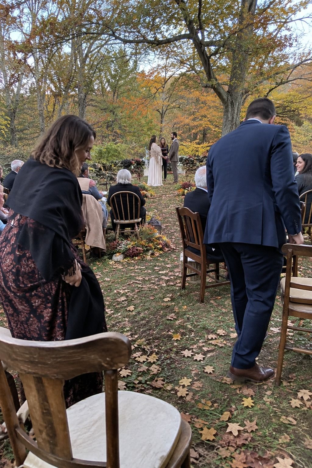 A guest arrives while two people take seats around a short curved aisle in a copper-leaf woodland clearing.