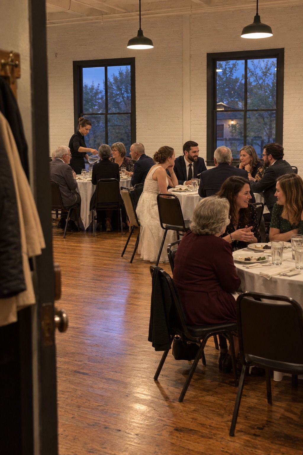 Mixed-age guests talk easily at four modest round tables while water is refilled in a flame-free community hall.