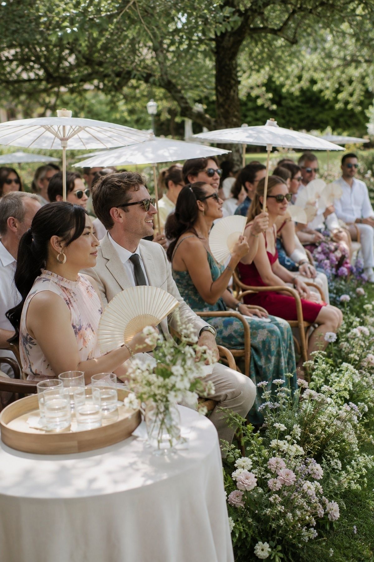 June ceremony guests sitting in shade with paper fans, white parasols, water, and garden flowers