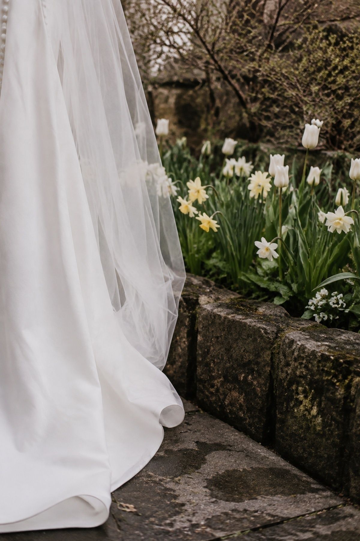 March wedding dress hem and veil photographed near tulips and daffodils beside a damp stone edge