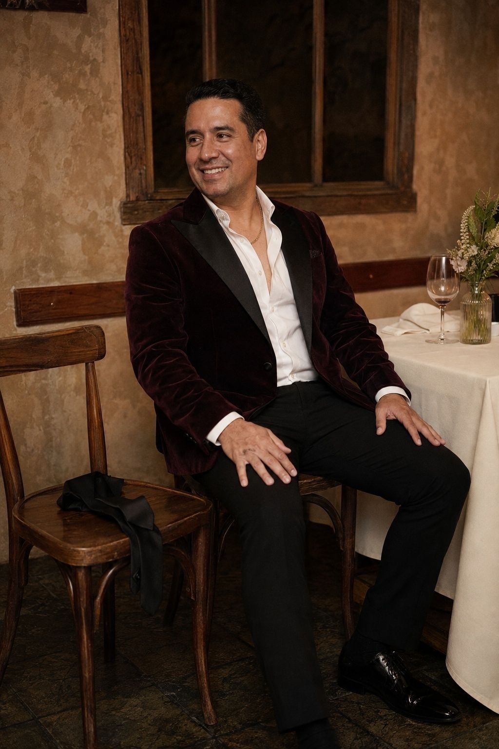 Groom seated at restaurant reception wearing a chain and open shirt after removing the tie, tie beside him