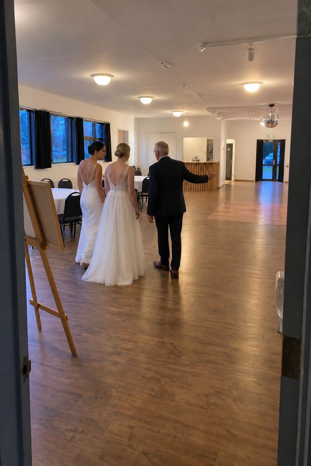 Two brides and a coordinator walk through separate entry, dining, bar, dance-floor, and exit zones in a restrained community hall.