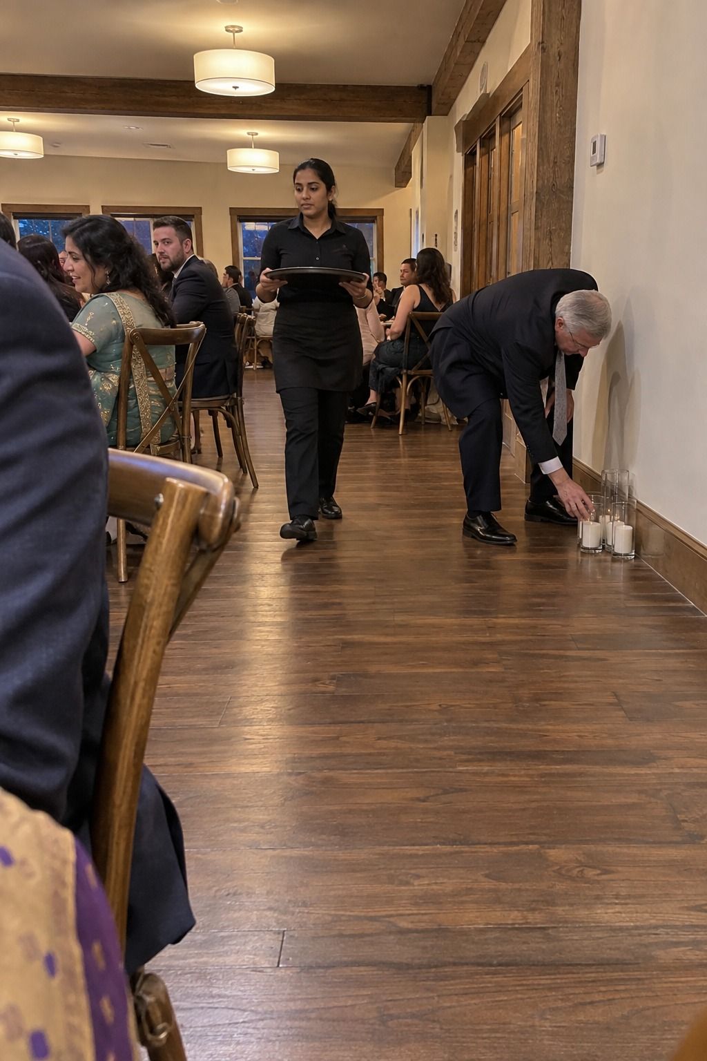 A server walks a wide open aisle while a coordinator moves low unlit glass holders fully against the wall.