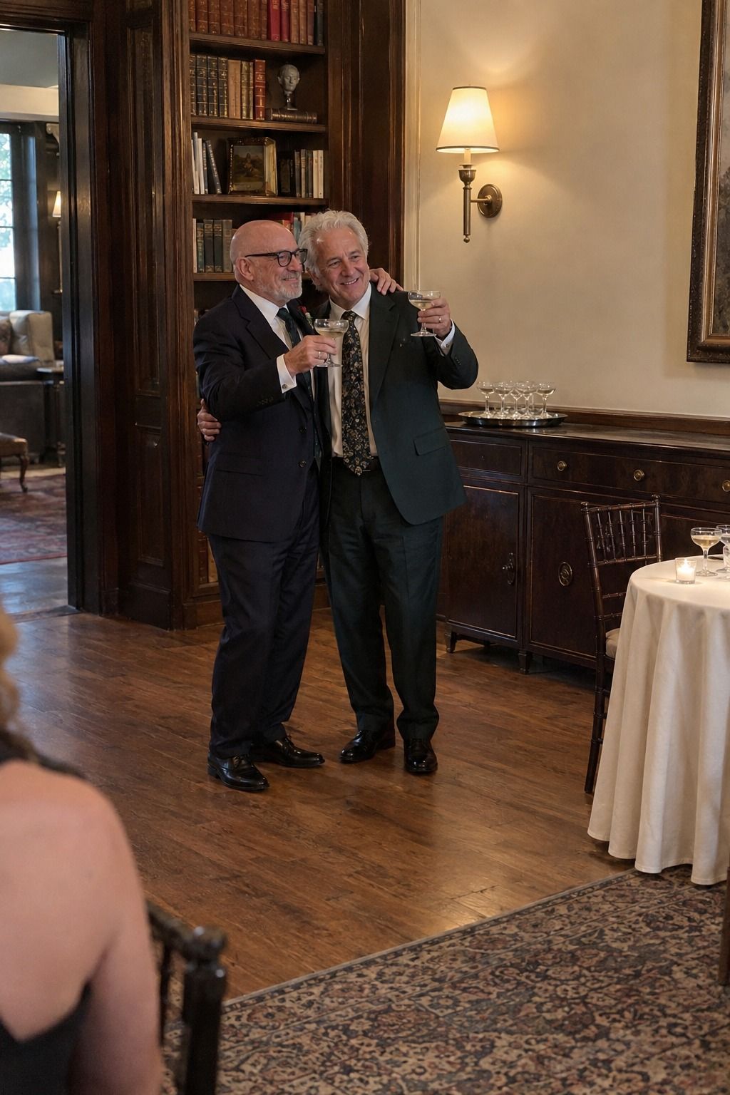 An older male couple hug and raise glasses in a breathable open spot with a secure tray and warm wall lamp nearby.