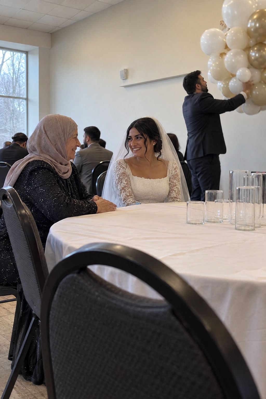 A bride and grandmother see each other clearly while a friend actively lifts an oversized balloon cluster away from their table.