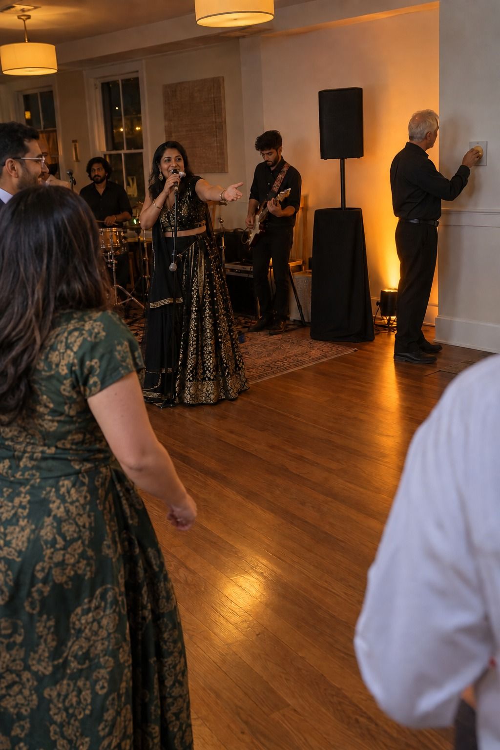 A bandleader invites guests closer while a technician turns a dimmer and a restrained amber wash defines the clear dance-floor edge.